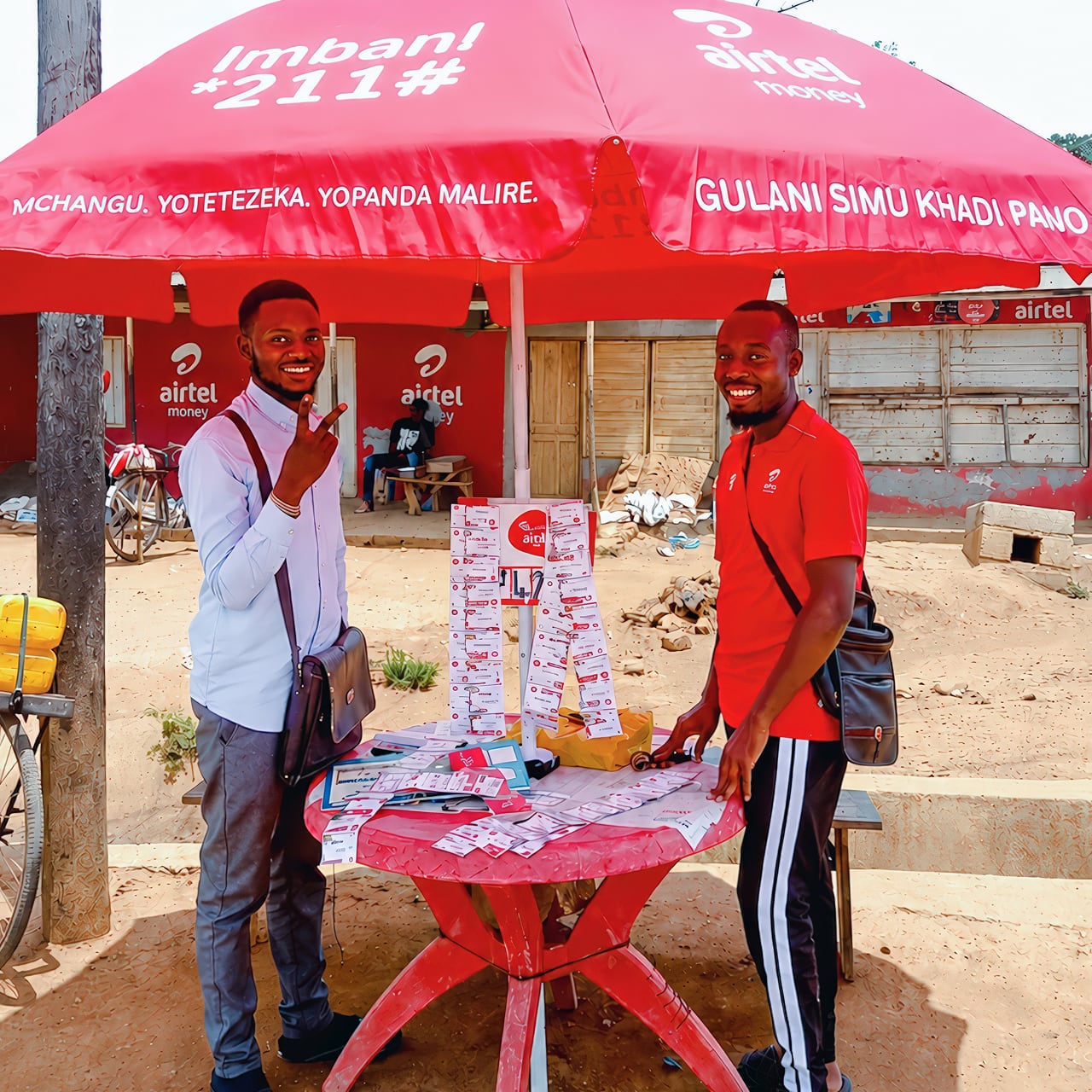 Two men standing either side of a table selling airtel africa products covered by a branded parasol