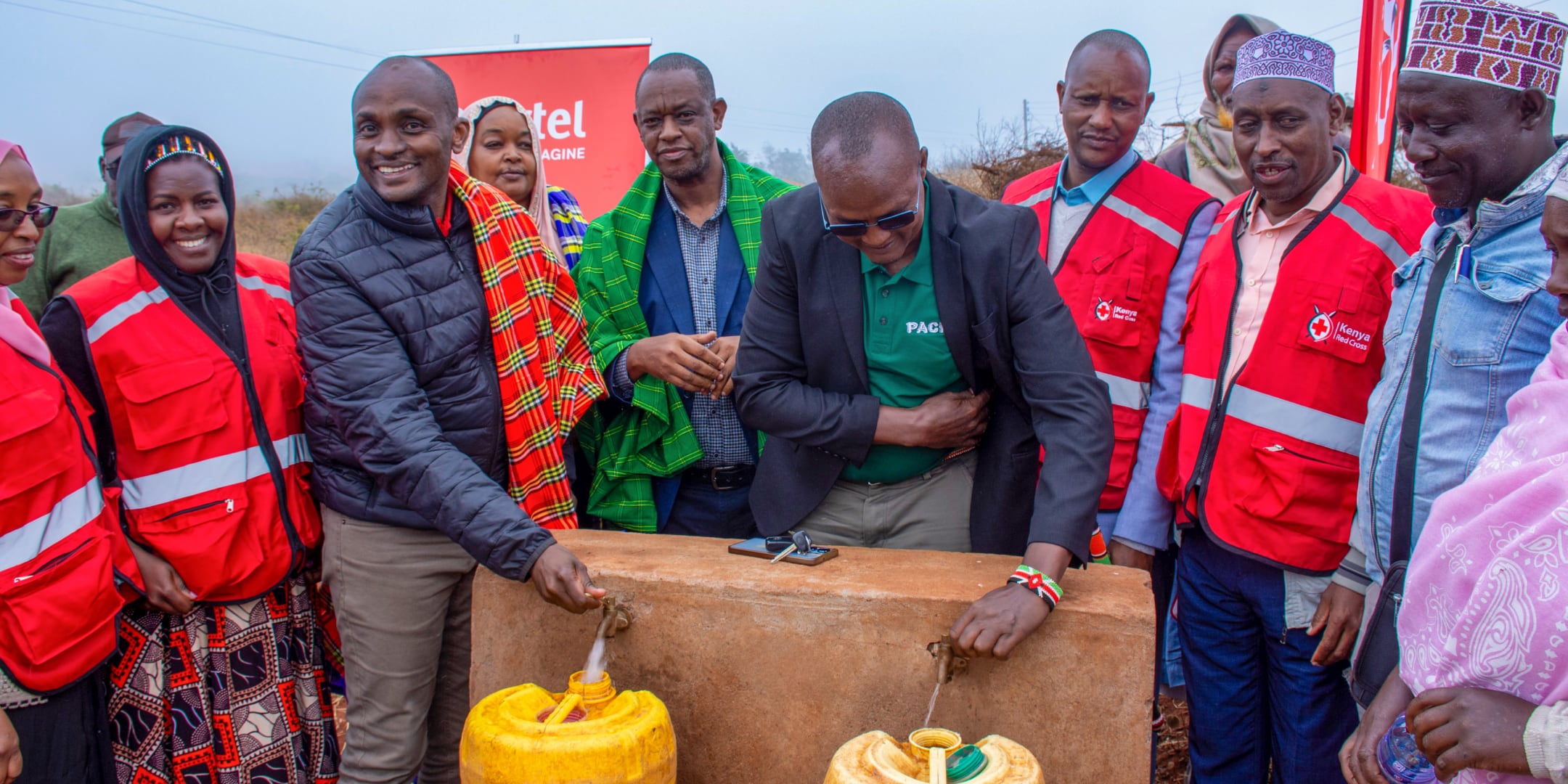 A group of people, some in red cross vests, standing around two open-air water tabs while filling up plastic containers