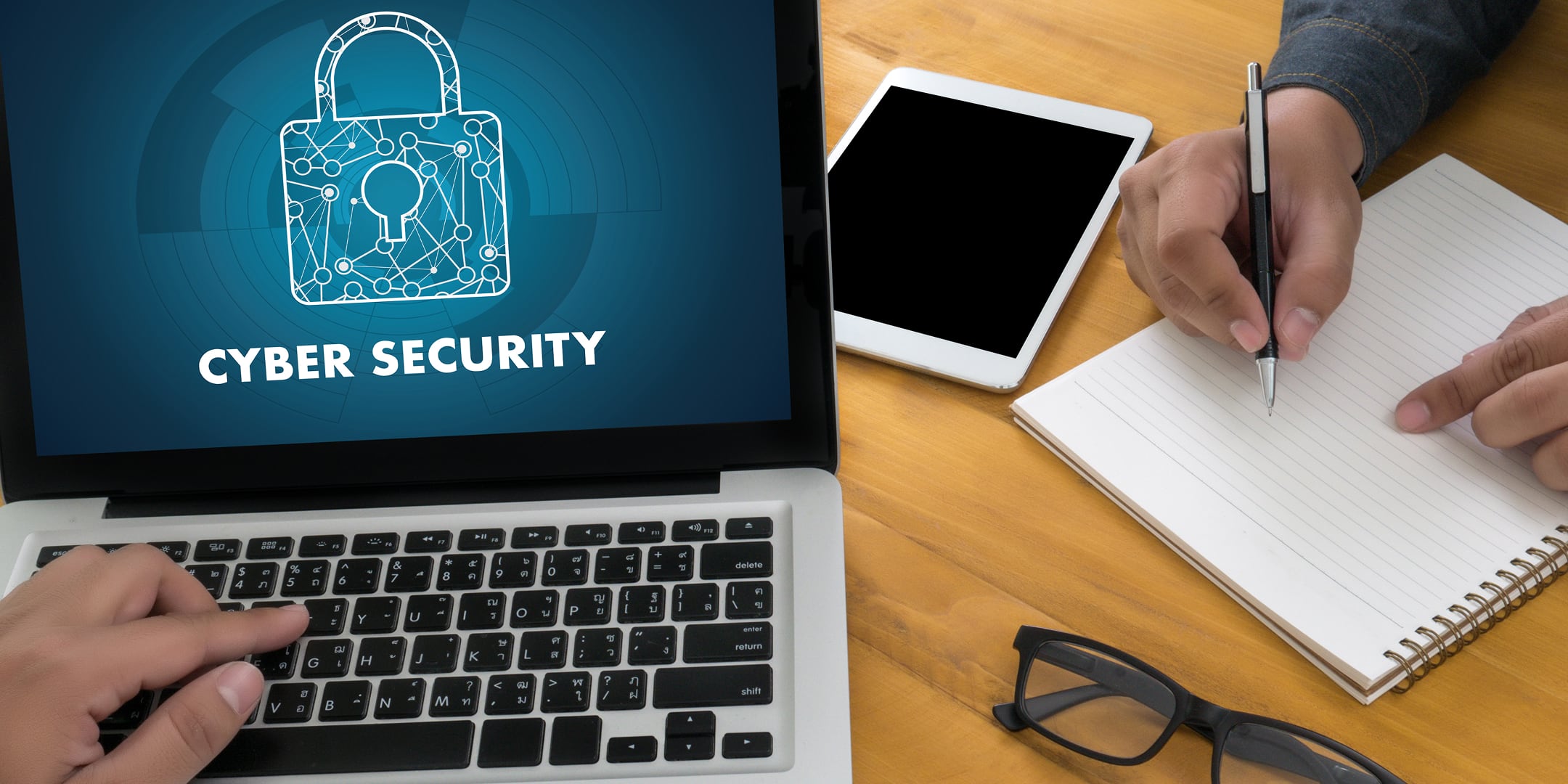 A close up of a laptop screen displaying the word 'cyber security' while a person opposite the user takes notes on a pad