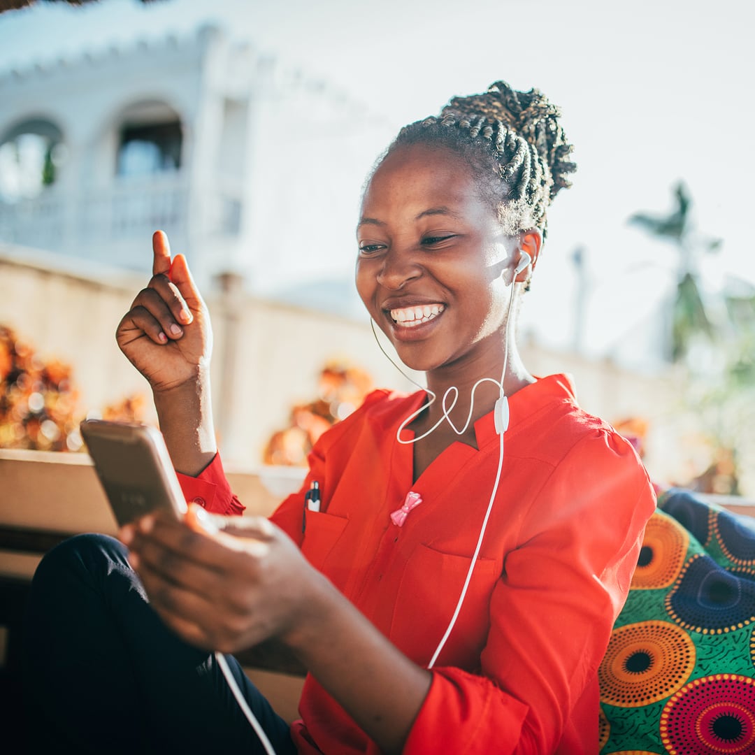 A woman sitting outside with headphones plugged into her ears and animatedly smiling at the phone in her hand