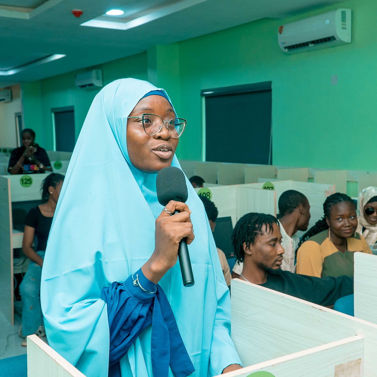 A young woman standing up and talking into a microphone while other students sit at desks in the background