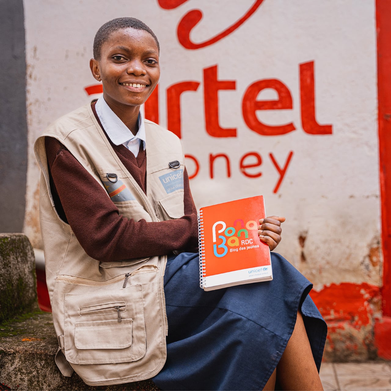 A young woman sitting in front of an Airtel Money mural holding up a UNICEF booklet