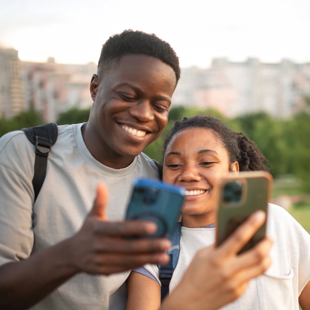 A young woman and man wearing backpacks are looking at their phones and smiling together in front of a blurred park and cityscape