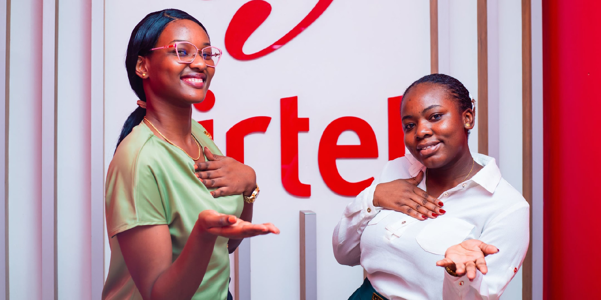 Two women in front of a wall displaying the Airtel logo each placing one hand on their chest and the open palm of the other hand towards the viewer